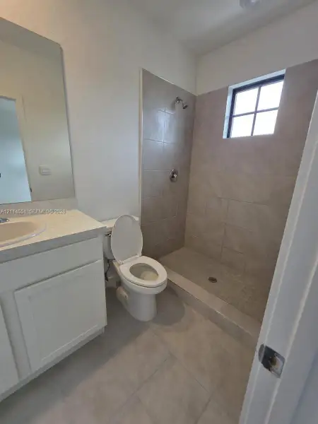 Bathroom featuring a vanity with a light-toned countertop, a wall-mounted mirror, a porcelain toilet, and a walk-in shower with tile surround and a window