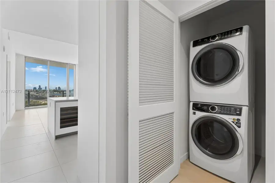 Laundry closet featuring stacked front-load washer and dryer with louvered door, neutral tile flooring, and built-in kitchen island with under-counter wine refrigerator
