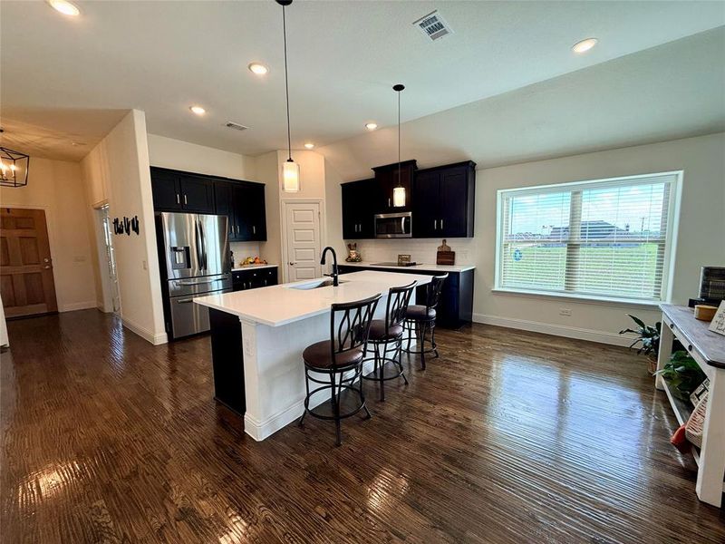 Open-concept kitchen featuring an expansive central island with seating, dark wood-finish flooring, white countertops, dark cabinetry, and recessed lighting Open-concept kitchen featuring an expansive central island with seating, dark wood-finish flooring, white countertops, dark cabinetry, and recessed lighting