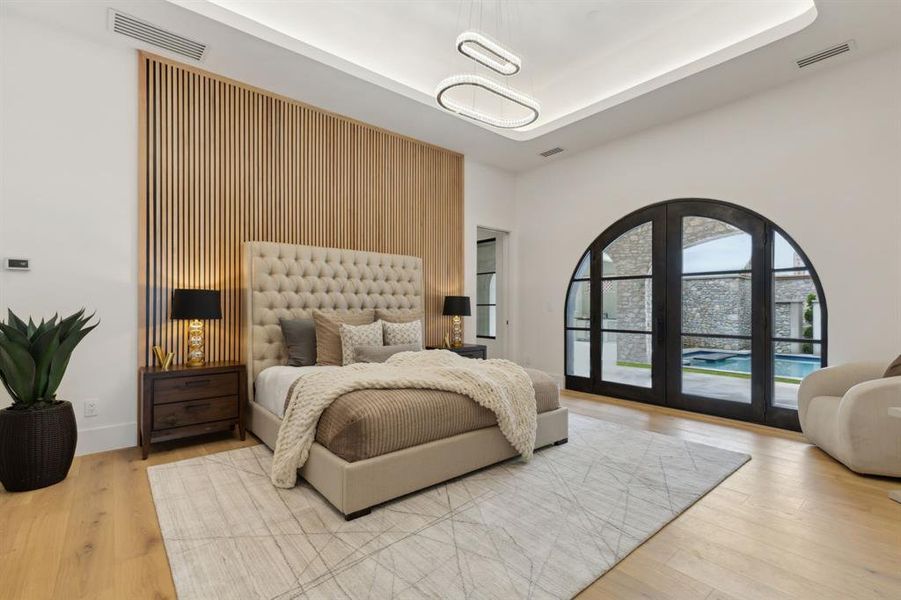 Bedroom with access to courtyard through iron and glass French doors , designer lighting, a tray ceiling, white oak wood finished floors, a linear fireplace and wood-paneled accent wall Bedroom with access to courtyard through iron and glass French doors , designer lighting, a tray ceiling, white oak wood finished floors, a linear fireplace and wood-paneled accent wall