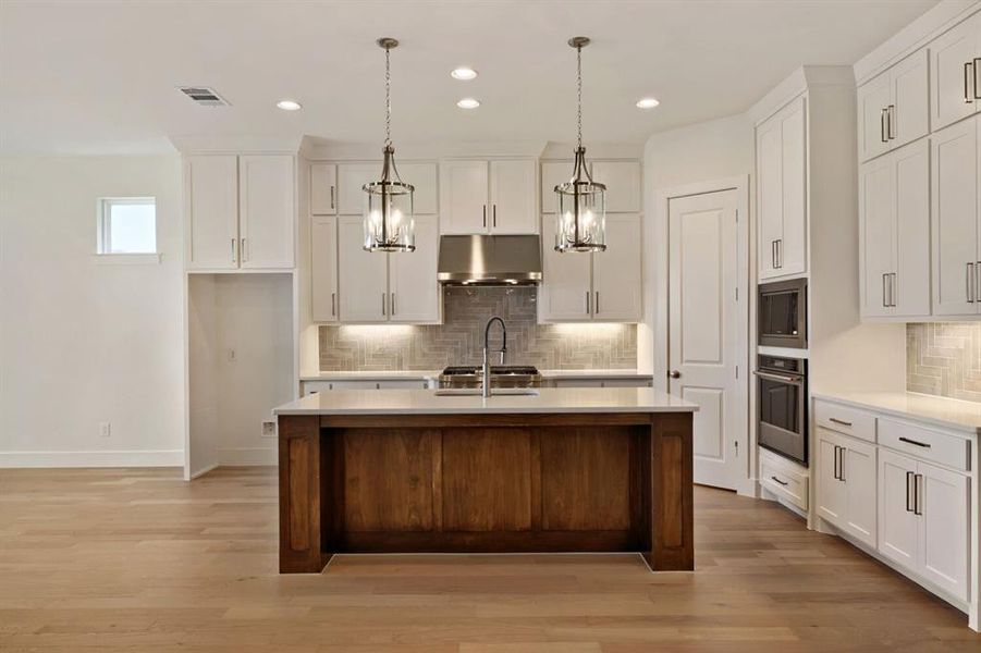 Two tone kitchen featuring a center island with sink, decorative backsplash, stainless steel appliances, hanging light fixtures, and light wood-style flooring