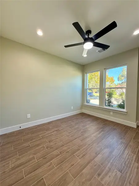 This room is bright and airy with large windows, wood-look tile flooring, and recessed lighting. It features a modern ceiling fan and neutral walls, creating a fresh and inviting space.