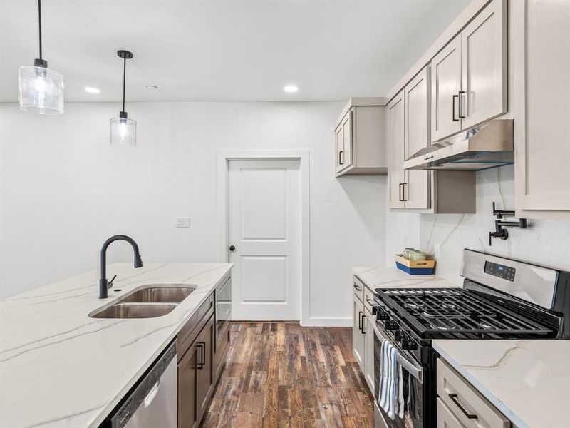 Kitchen featuring appliances with stainless steel finishes, light stone counters, dark wood-type flooring, under cabinet range hood, and decorative light fixtures