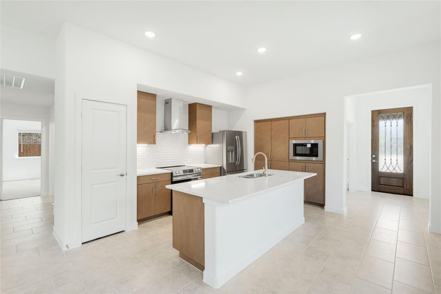 Open kitchen with wood cabinets, white island with sink, and stainless steel appliances on light tile floors