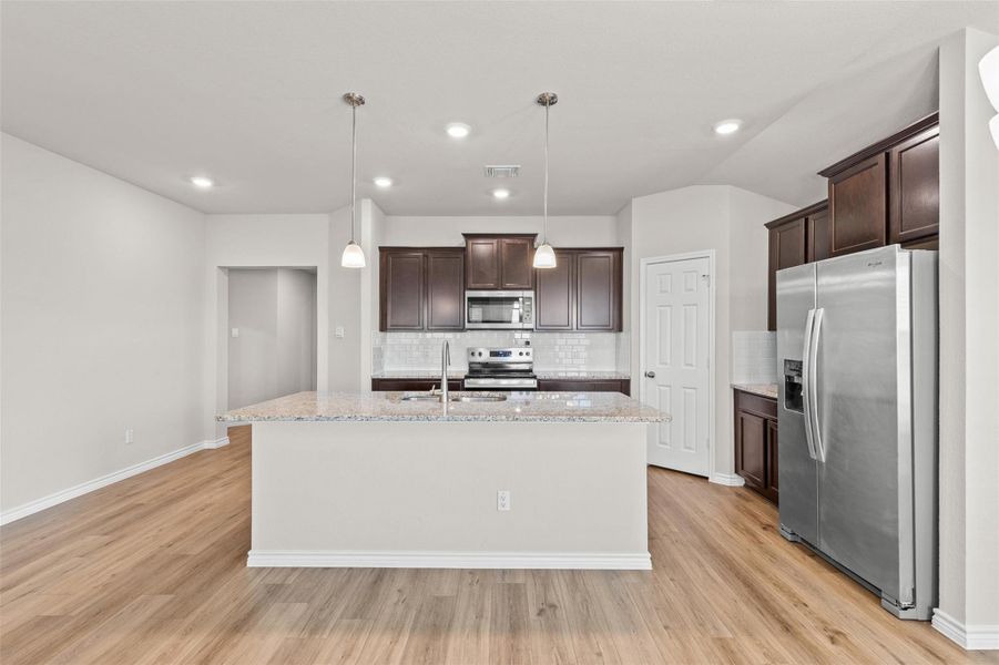 Kitchen with stainless steel appliances, dark wood finish cabinetry, light stone counters, and an island with sink