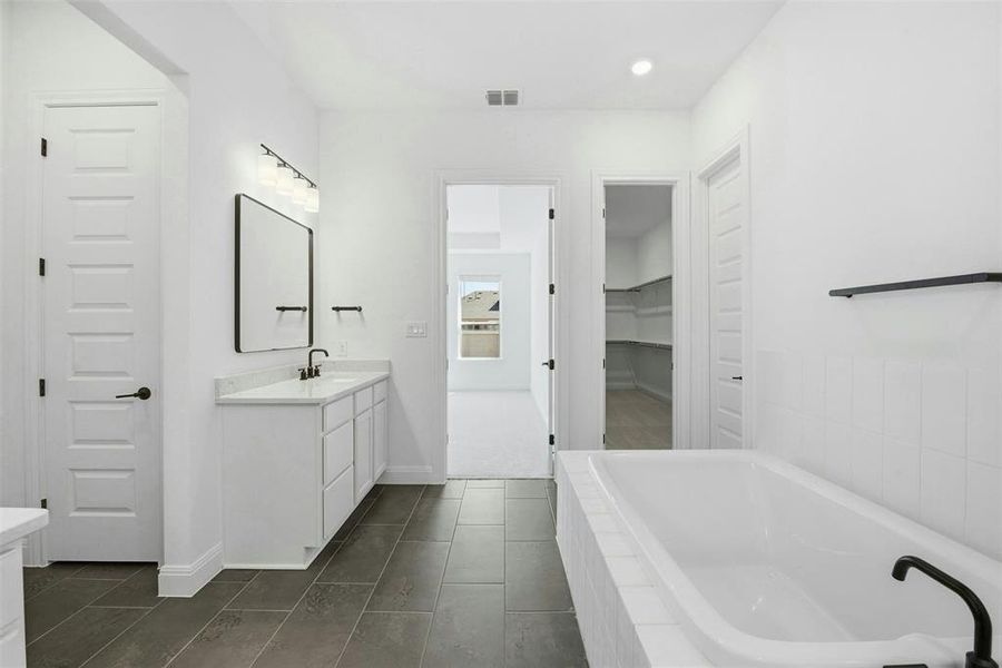Spacious bathroom featuring dark tile flooring, a built-in soaking tub with matte black fixtures, a double vanity with a light-toned countertop, and a large framed mirror with overhead lighting