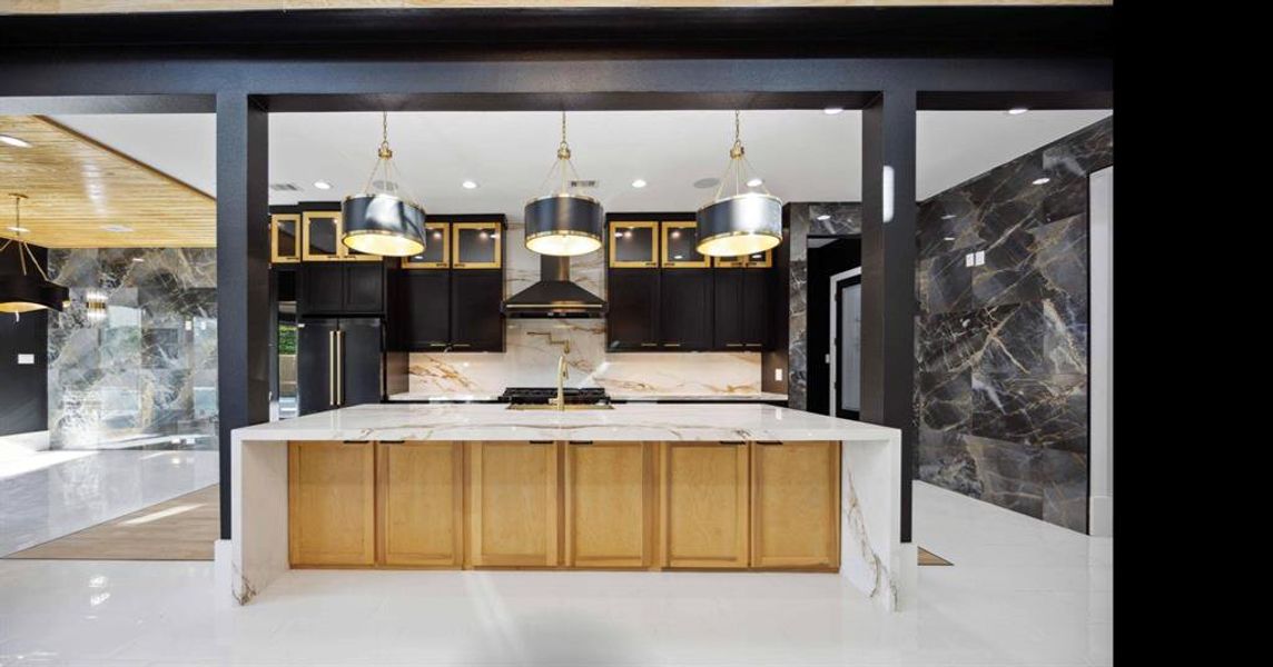 This is a modern kitchen featuring a large central island with a marble countertop, sleek black cabinetry with gold accents, and stylish pendant lighting. The kitchen is framed by dark, bold architectural elements and complemented by luxurious marble wall finishes.