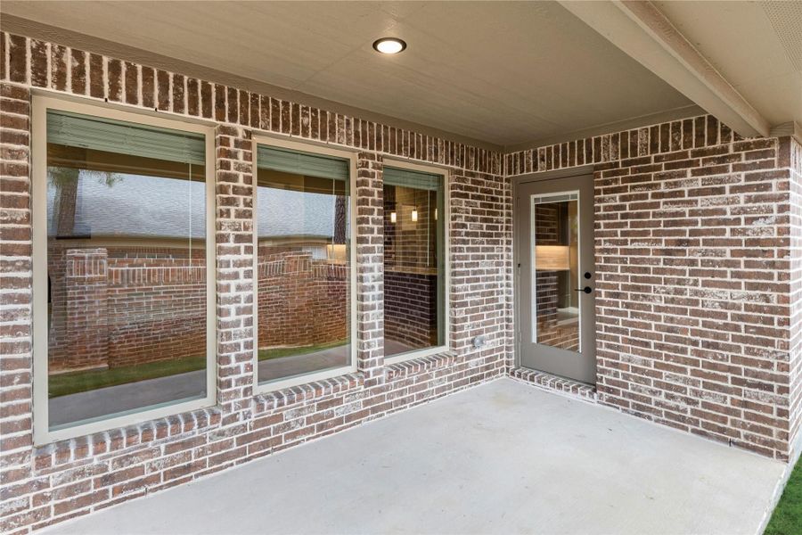 Covered back patio with concrete floor, brick exterior walls, large windows, and a glass back door with overhead light.