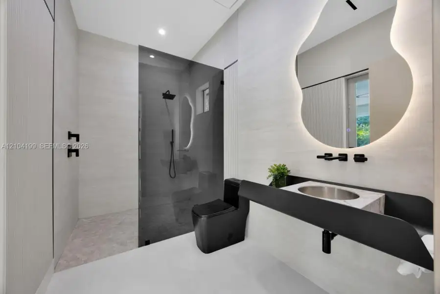 Bathroom featuring a wall-mounted floating vanity with a stone countertop, backlit organic-shaped mirror, matte black fixtures, and a walk-in shower with floor-to-ceiling tile