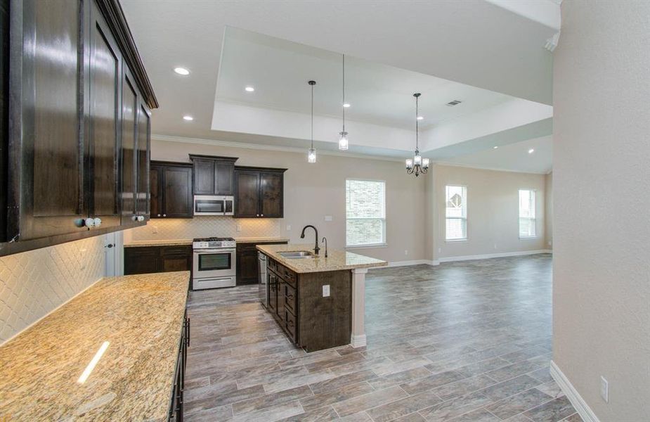 This is a spacious, open-concept kitchen and living area featuring dark wood cabinetry, stainless steel appliances, granite countertops, and modern pendant lighting. The room is well-lit with natural light from multiple windows and offers versatile space for dining and relaxation.