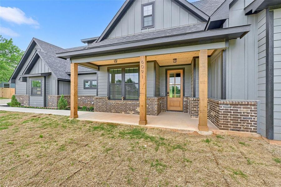 View of exterior entry with brick siding, board and batten siding, roof with shingles, and covered porch
