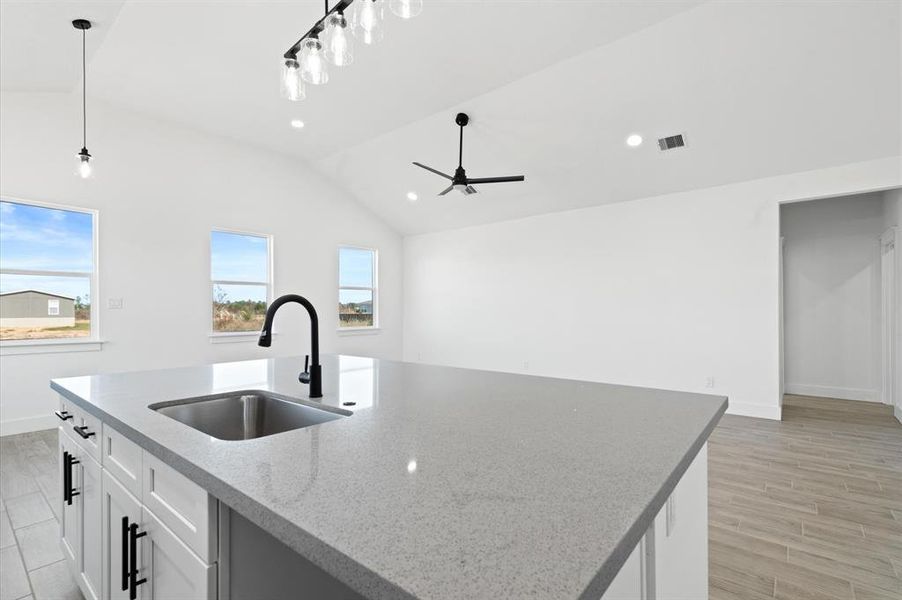 This is a spacious, modern kitchen featuring a large island with a sink and high faucet, contemporary lighting fixtures, and ample natural light from several windows. It has a clean, minimalist design with white walls and light wood flooring.