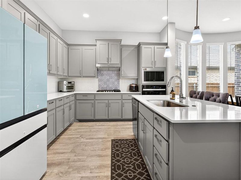 Modern kitchen featuring light gray cabinetry, light countertops, wood-finish flooring, a patterned tile backsplash, and a central island with a sink Modern kitchen featuring light gray cabinetry, light countertops, wood-finish flooring, a patterned tile backsplash, and a central island with a sink