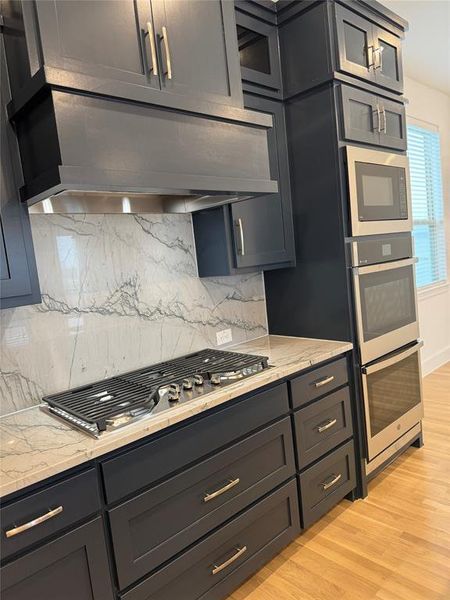 Kitchen with glass insert cabinets, appliances with stainless steel finishes, light wood-type flooring, and light stone counters