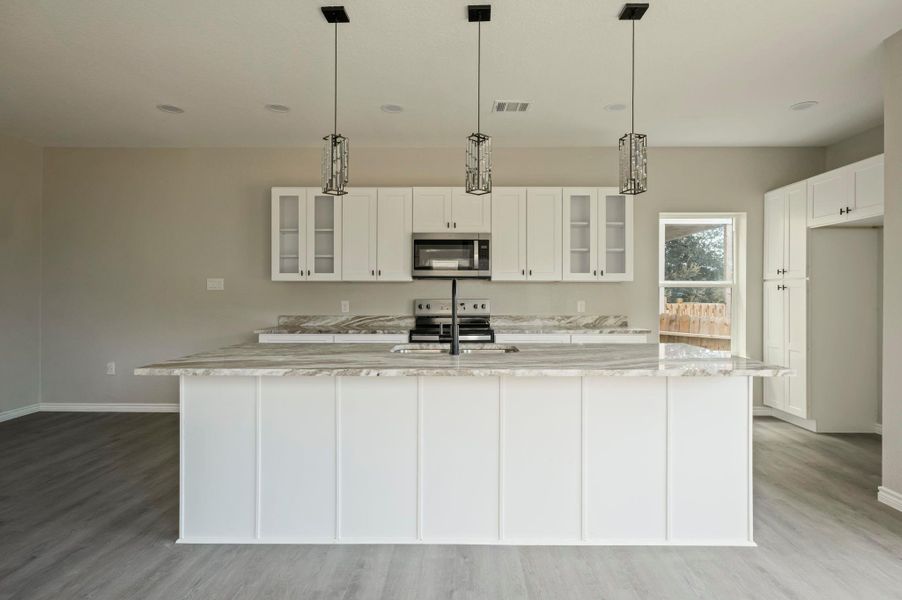 Kitchen with glass insert cabinets, light stone countertops, a center island with sink, white cabinets, and hanging light fixtures