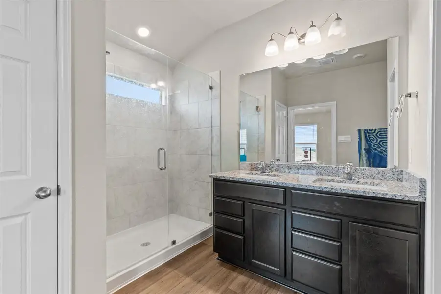 Bathroom featuring a stall shower, double vanity, light wood-style flooring, and vaulted ceiling