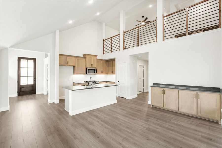 Open-concept kitchen with high ceilings, center island, light wood cabinets, and hardwood floors overlooking a second-floor loft railing. Open-concept kitchen with high ceilings, center island, light wood cabinets, and hardwood floors overlooking a second-floor loft railing.