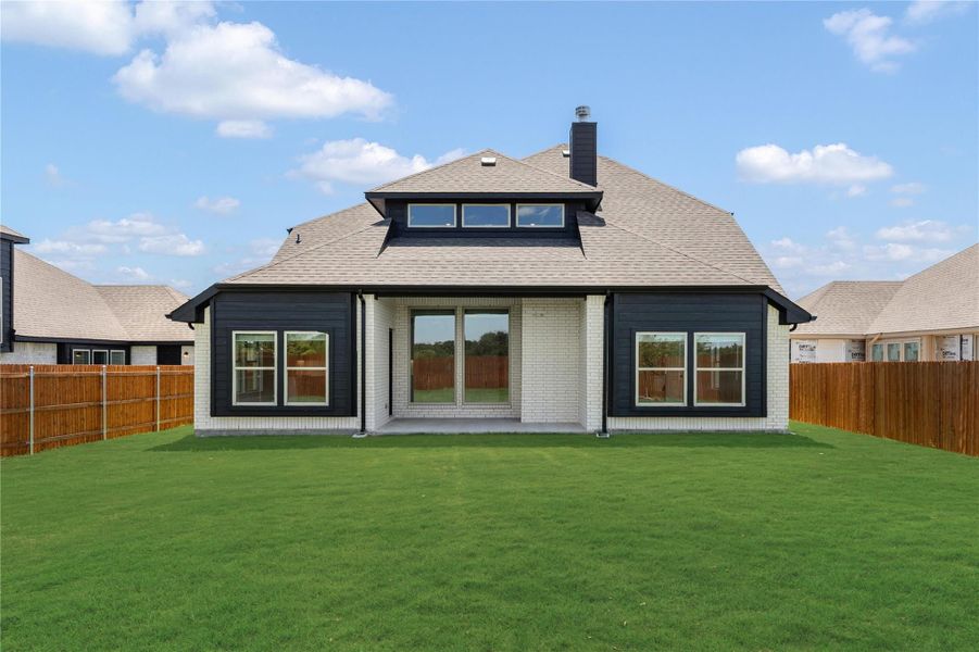Rear view of home with dark siding, white brick, covered patio, and fenced backyard with green lawn