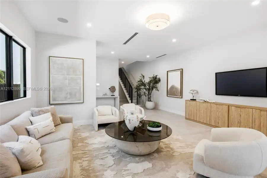 Open-concept living space featuring neutral oversized tile flooring, recessed ceiling lighting, a contemporary flush-mount fixture, black-framed windows, and built-in wood-finish cabinetry
