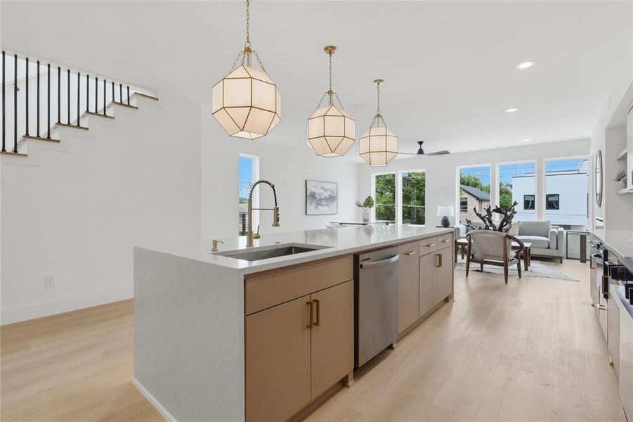 Open-concept kitchen and living area featuring a large center island with integrated sink, light-toned wood-finish flooring, abundant natural light from multiple windows, modern pendant lighting, and a contemporary staircase with black railing Open-concept kitchen and living area featuring a large center island with integrated sink, light-toned wood-finish flooring, abundant natural light from multiple windows, modern pendant lighting, and a contemporary staircase with black railing