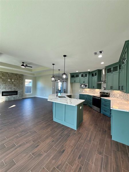 This open-concept space features a modern kitchen with teal cabinets, marble countertops, and stylish pendant lighting. It seamlessly connects to a living area with wood-look tile flooring, a stone fireplace, and ample natural light from large windows. Ideal for entertaining and family gatherings. This open-concept space features a modern kitchen with teal cabinets, marble countertops, and stylish pendant lighting. It seamlessly connects to a living area with wood-look tile flooring, a stone fireplace, and ample natural light from large windows. Ideal for entertaining and family gatherings.