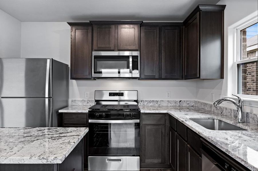 Kitchen featuring stainless steel appliances, light stone countertops, dark brown cabinets, and sink