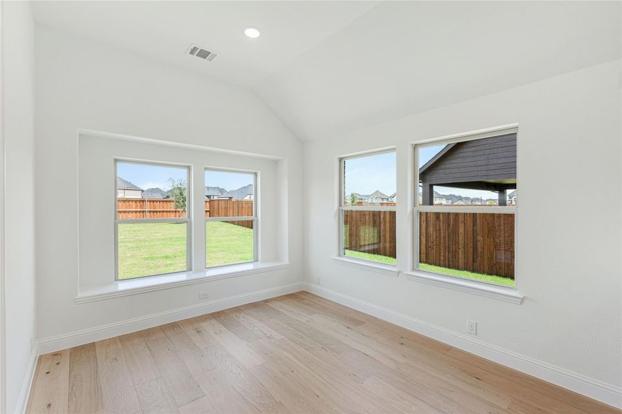 Empty room with vaulted ceiling, light hardwood floors, and four windows overlooking a fenced backyard