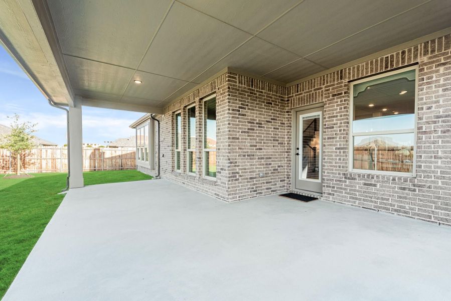Covered back patio with concrete slab, brick exterior, back door, and fenced yard with green lawn
