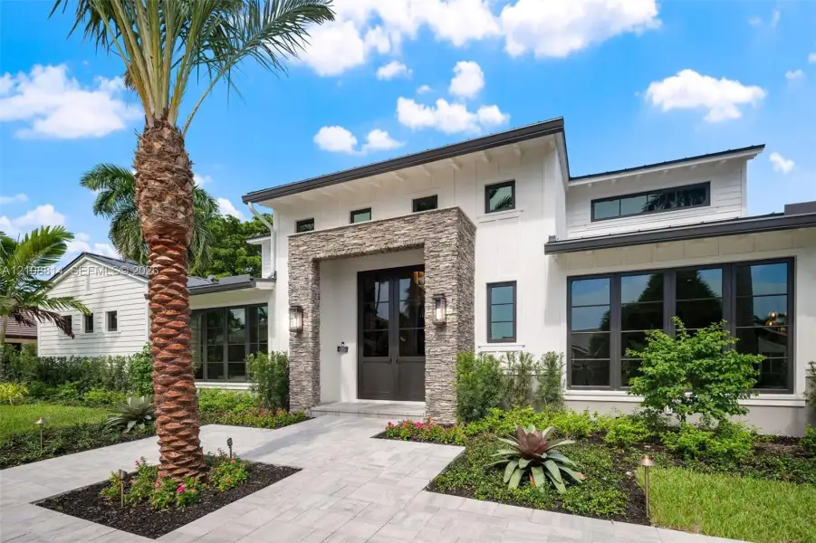Modern exterior featuring white vertical siding, a stone veneer entryway surround, metal roof accents, expansive dark-framed windows, and a paved walkway Modern exterior featuring white vertical siding, a stone veneer entryway surround, metal roof accents, expansive dark-framed windows, and a paved walkway