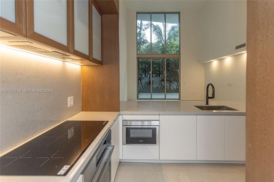 Kitchen featuring custom wood-toned cabinetry with glass-paneled upper doors, stone countertops, a flat-surface induction cooktop, integrated stainless steel appliances, and floor-to-ceiling windows