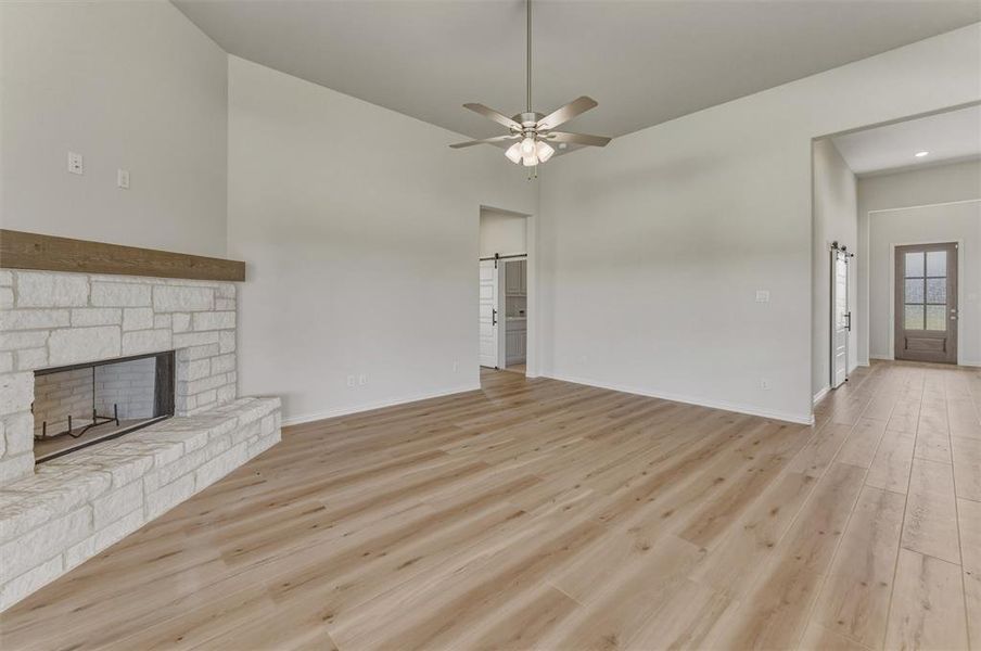 Spacious living area featuring light wood-finish flooring, a stone fireplace with a wood mantle, a ceiling fan, vaulted ceilings, and white interior walls Spacious living area featuring light wood-finish flooring, a stone fireplace with a wood mantle, a ceiling fan, vaulted ceilings, and white interior walls