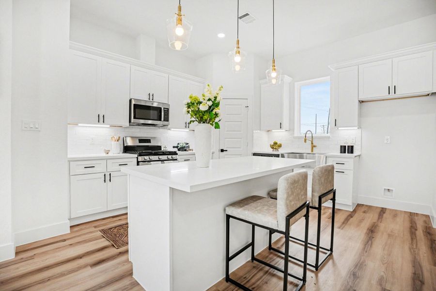 This modern kitchen features sleek white cabinetry, stainless steel appliances, and a spacious island with seating. Pendant lighting adds elegance, while wood flooring provides warmth and style.