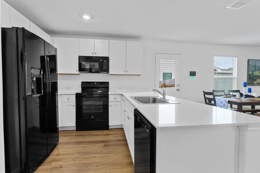 Modern kitchen featuring wood-finish flooring, white cabinetry, integrated black appliances, a central island with a stainless steel sink, and white countertops