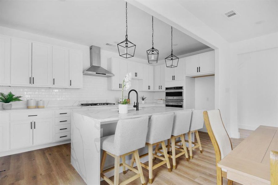 Kitchen featuring white cabinetry, wall chimney range hood, light hardwood / wood-style flooring, decorative light fixtures, and a center island with sink