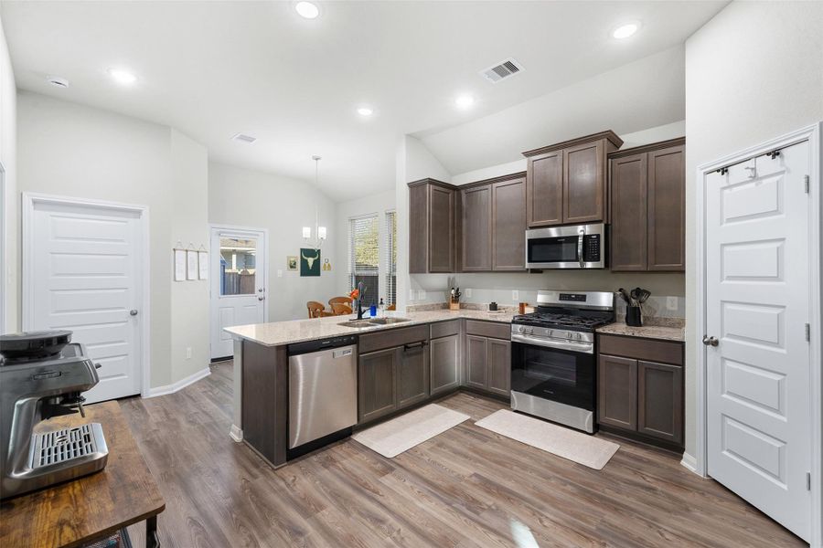 This kitchen features modern stainless steel appliances, dark wood cabinetry, and granite countertops. It has an open layout with ample natural light and a dining area visible in the background.