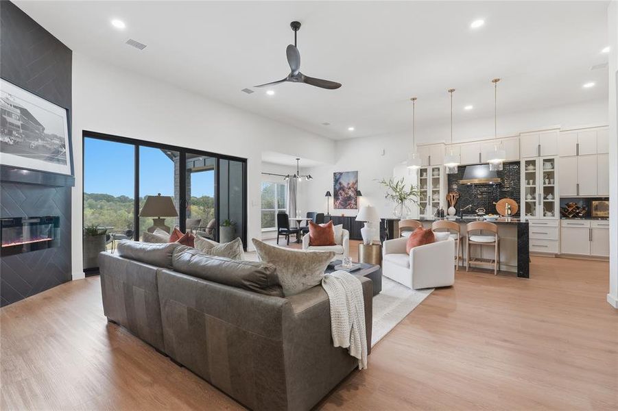 Open-concept living space featuring wood-finish flooring, a contemporary fireplace with a dark herringbone tile surround, and expansive glass doors opening to outdoor scenery
