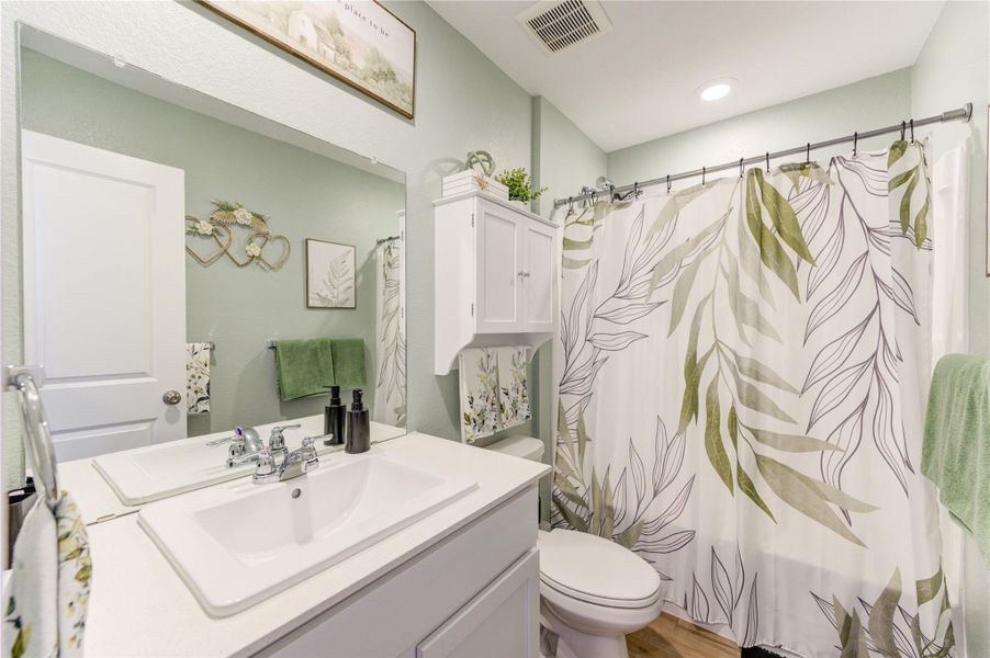 This is a modern bathroom featuring a sleek white vanity with dual sinks, a stylish mirror, and ample storage. The shower has a leafy-patterned curtain, complemented by light green walls and wood look alike-style flooring, creating a fresh, inviting atmosphere.