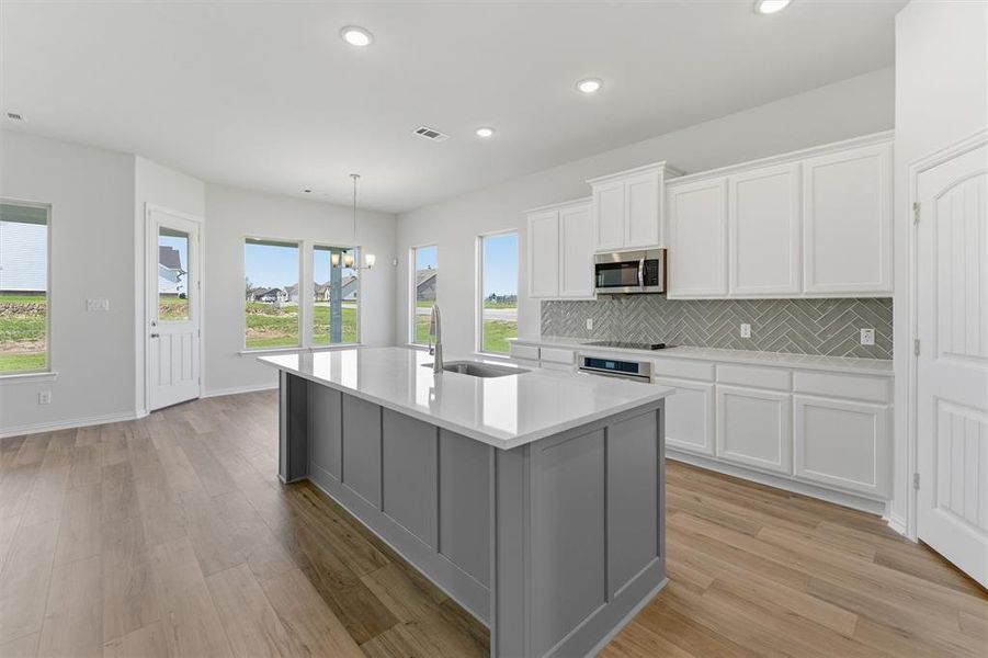 Open-concept kitchen featuring a large island with a sink, white cabinetry, integrated stainless steel appliances, a herringbone tile backsplash, and light wood-finish flooring