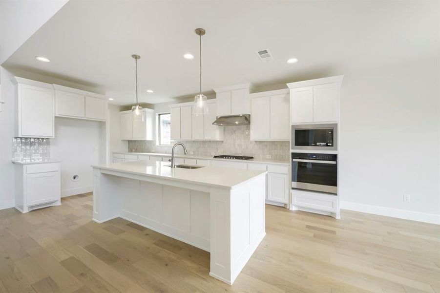 Spacious kitchen featuring an expansive central island, light wood-finish flooring, white cabinetry, integrated stainless steel appliances, and a tiled backsplash Spacious kitchen featuring an expansive central island, light wood-finish flooring, white cabinetry, integrated stainless steel appliances, and a tiled backsplash