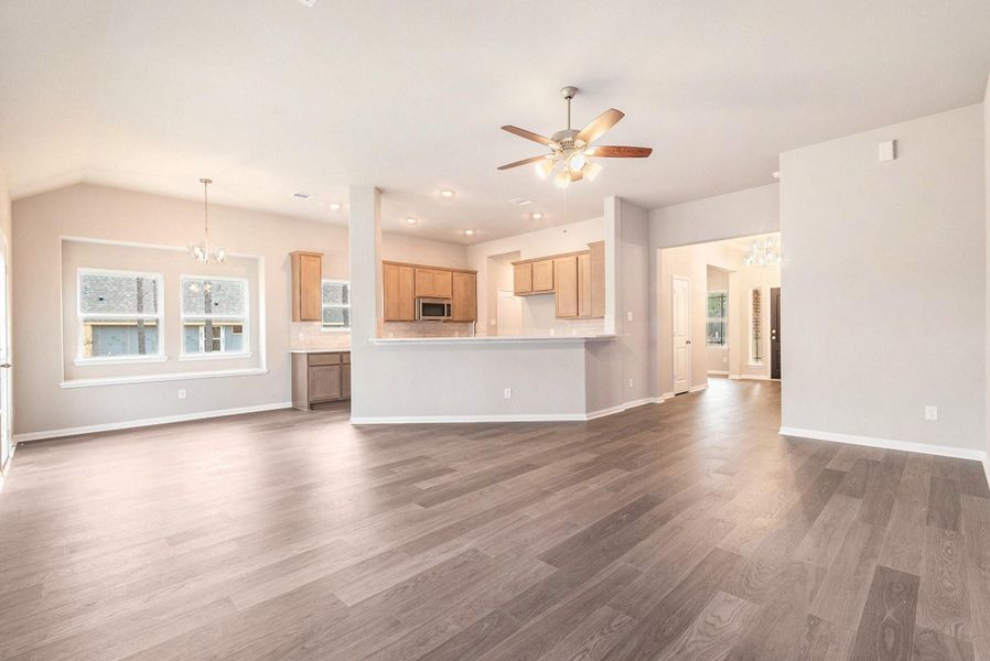 This spacious open-concept living area features light wood flooring, neutral walls, and ample natural light. The modern kitchen with light wood cabinets and a breakfast bar seamlessly connects to the dining and living spaces. A ceiling fan and recessed lighting enhance the airy feel, while large windows provide a view of the backyard.