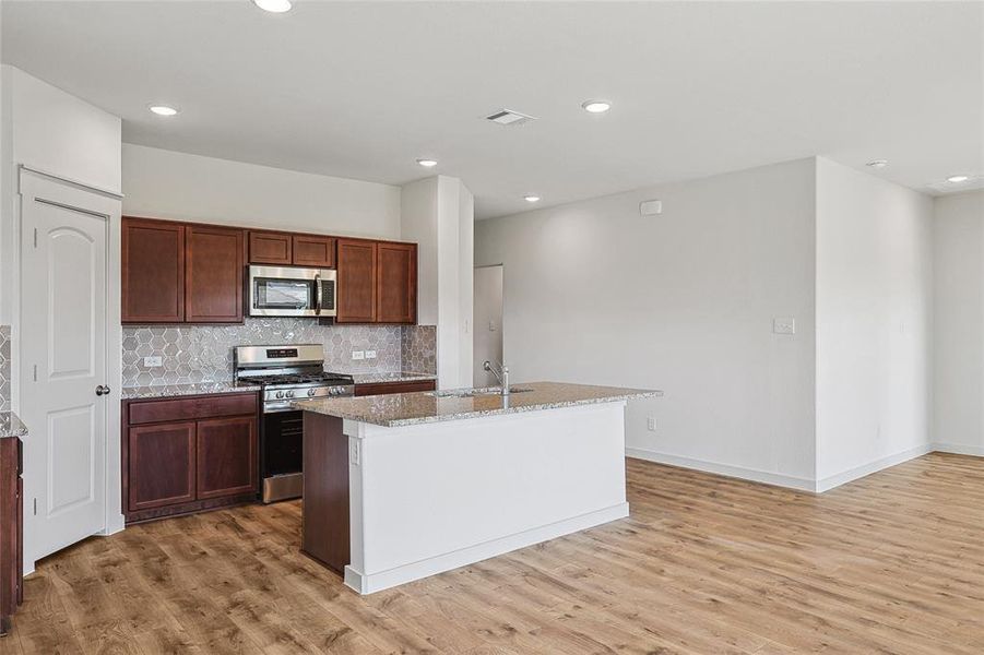 Kitchen featuring light hardwood / wood-style floors, tasteful backsplash, light stone counters, an island with sink, and appliances with stainless steel finishes