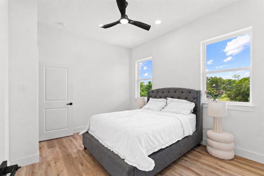 Light wood-finish flooring complements the bright white walls of this bedroom, featuring two large windows, a white paneled door with black hardware, and a modern ceiling fan with integrated lighting