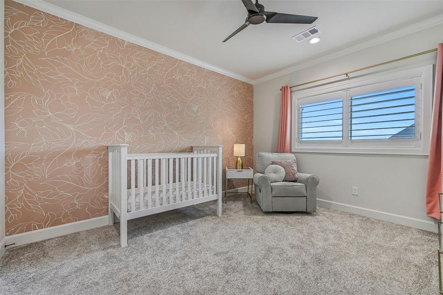 Bedroom two upstairs continues with plush carpet, crown molding, plantation shutters & a ceiling fan, while a soft floral accent wall adds personality to the generously sized space.
