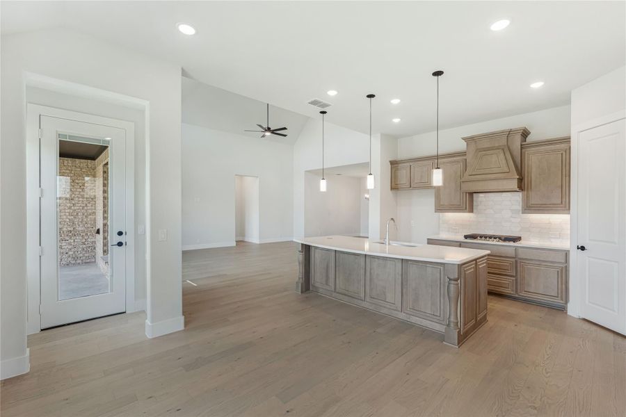 Open kitchen with large center island, tan wood cabinets, stone backsplash, and light hardwood floors Open kitchen with large center island, tan wood cabinets, stone backsplash, and light hardwood floors