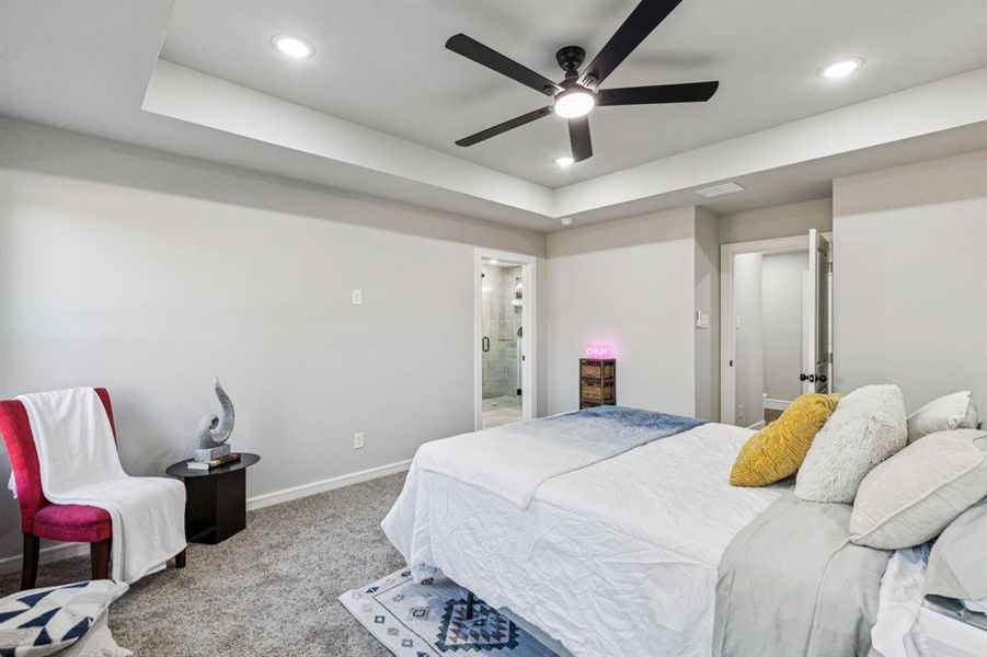 Carpeted bedroom with a ceiling fan, a tray ceiling, ensuite bathroom, and recessed lighting