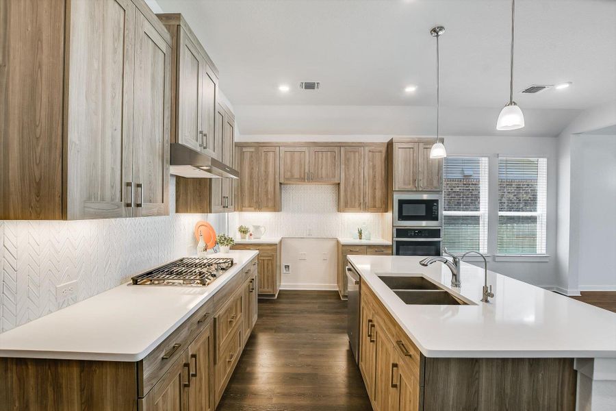 Modern kitchen featuring wood-finish cabinetry, white countertops, and a herringbone-patterned backsplash. Built -in oven and microwave with 5-buner gas cooktop.