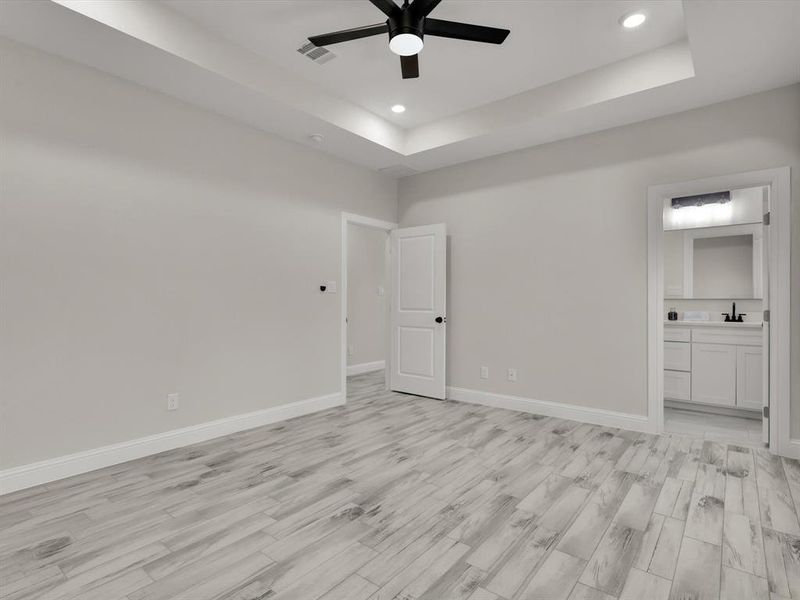 Unfurnished bedroom featuring a raised ceiling, light hardwood / wood-style flooring, ensuite bath, and ceiling fan