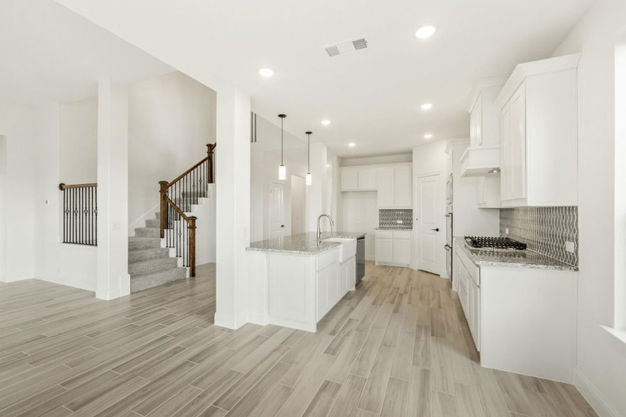 Open kitchen with white cabinets, granite island, tile backsplash, and wood-look floors, with staircase visible nearby. Open kitchen with white cabinets, granite island, tile backsplash, and wood-look floors, with staircase visible nearby.