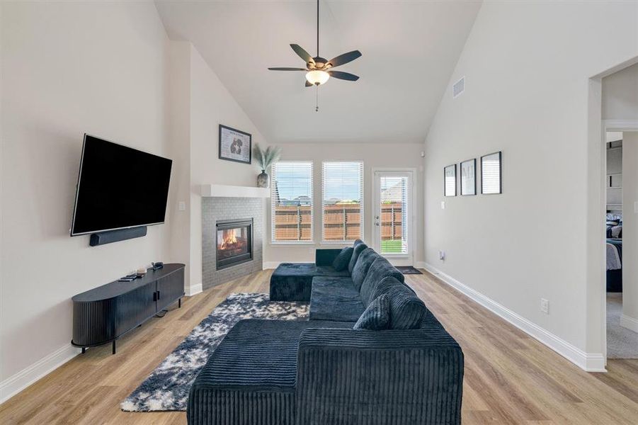 Vaulted living space featuring a fireplace with a textured tile surround and mantel, wood-finish flooring, a ceiling fan, and transom windows with backyard access