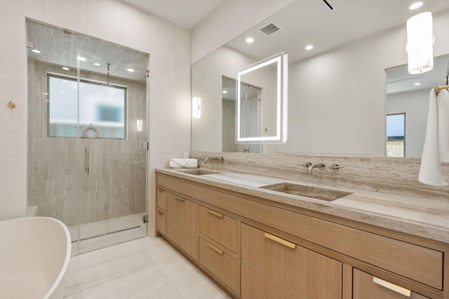 Spa-like primary bathroom showcasing a custom vanity, integrated makeup mirror, and chrome three-piece wall-mounted sink fixtures, with textured limestone detailing enhancing the shower exterior. Spa-like primary bathroom showcasing a custom vanity, integrated makeup mirror, and chrome three-piece wall-mounted sink fixtures, with textured limestone detailing enhancing the shower exterior.